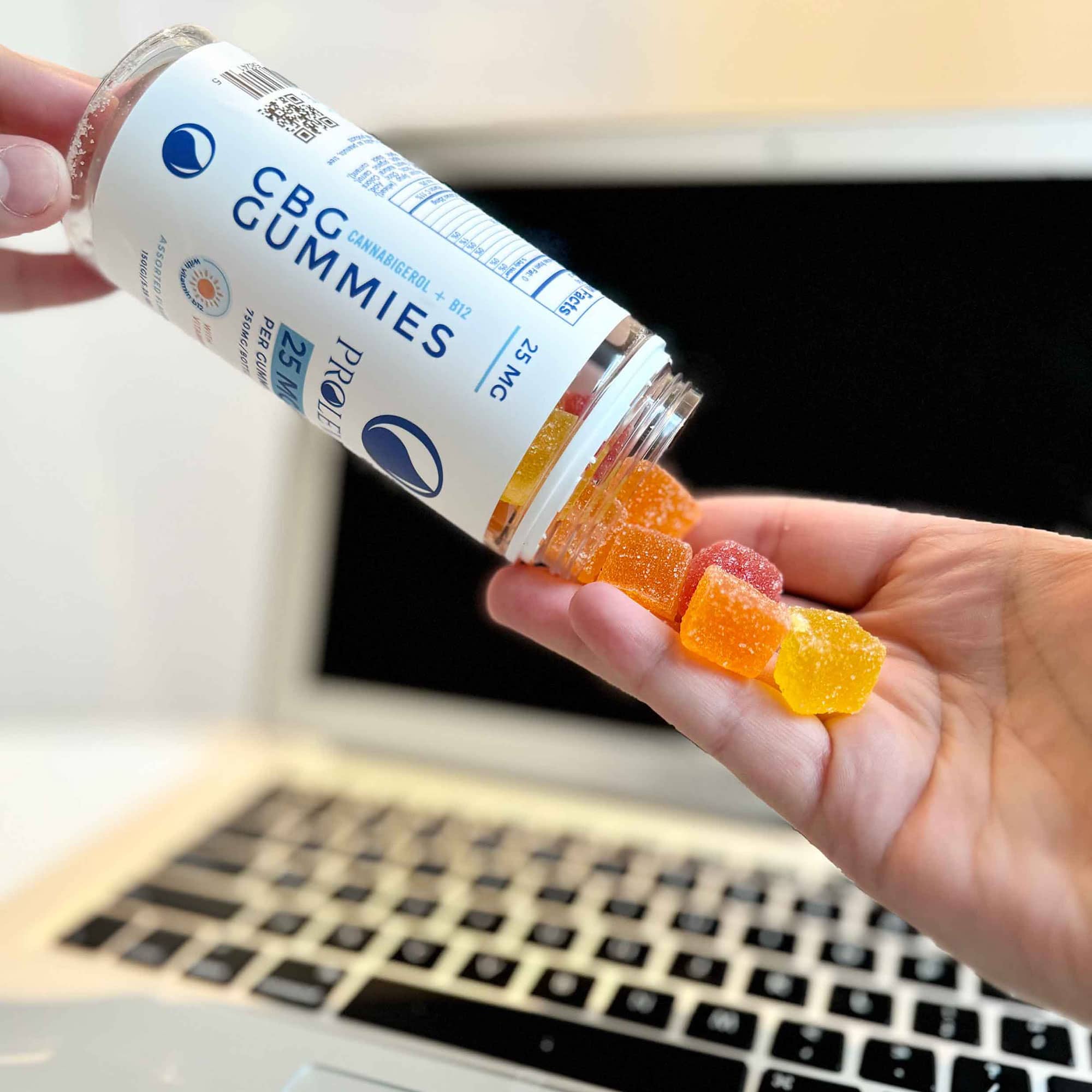 Image of CBG Gummies being poured into a hand from the bottle with a laptop in the backgroud